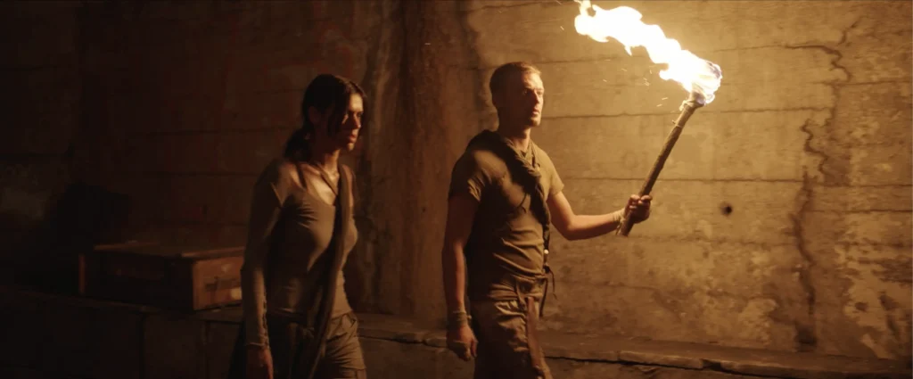 A man holding a burning torch walks through a dim underground tunnel followed by a woman, scene from the film Sanctuary